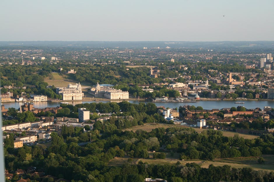 An aerial view of a cityscape featuring a wide river running through the middle, with historic and modern buildings lining the banks. In the foreground, there is a mix of green trees, parks, and residential areas with rooftops visible. The riverbanks are lined with trees and some boats are visible on the water. In the background, the city extends into a flat landscape with scattered buildings and patches of greenery, under a clear sky illuminated by soft daylight. This scene highlights urban and natural elements typical of a city with a historic skyline, which aligns with the context of managing and disposing of waste through private services such as those offered by Waste Disposal Greenwich, particularly in facilitating alternative rubbish removal solutions beyond traditional municipal collection.