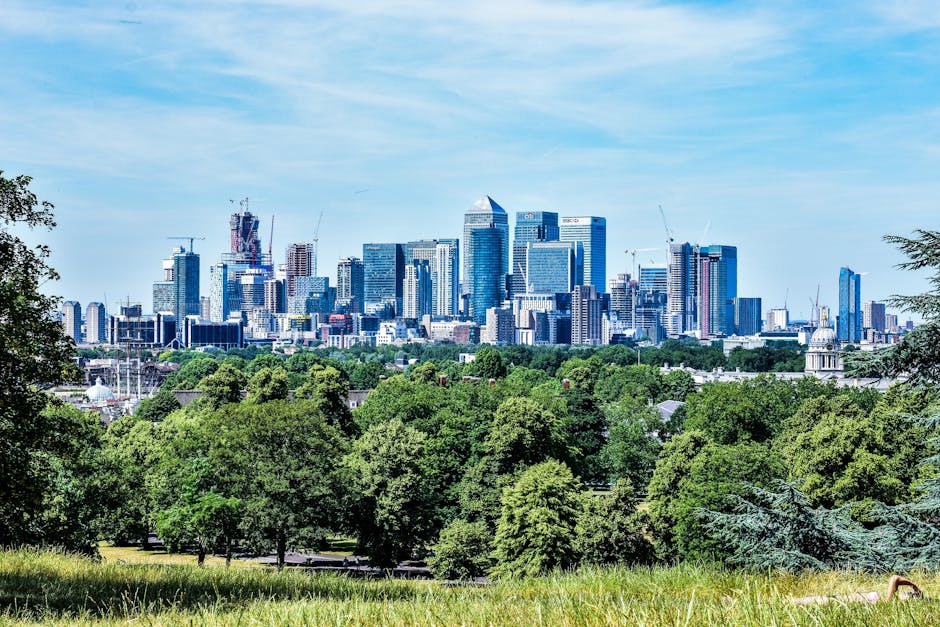 A panoramic view of a modern city skyline featuring numerous high-rise buildings with glass facades, some still under construction with visible cranes, set against a bright blue sky with scattered clouds. In the foreground, there is a lush green park with tall, leafy trees and a grassy area, providing a natural contrast to the urban environment. The scene is well-lit, indicating daytime, with clear visibility of the architectural details and vibrant greenery. The presence of the park suggests an area of open space within or near the city, possibly serving as a location for outdoor activities or relaxation, and highlights the mix of natural and built environments often seen in cityscapes where alternative waste management solutions might be considered for maintaining such green spaces.