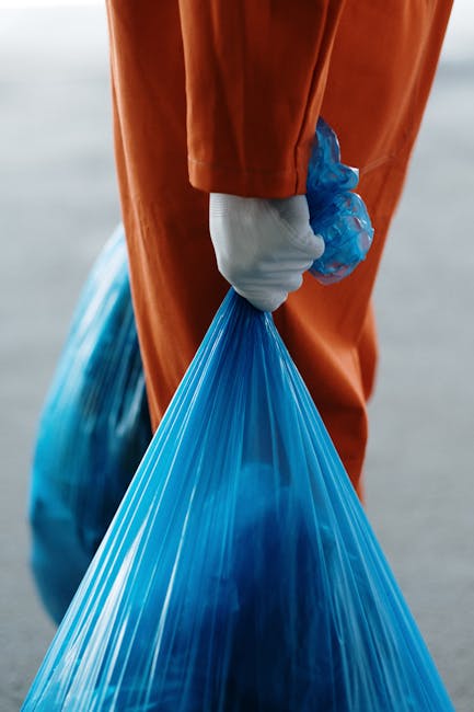 A close-up view of a person wearing an orange work jumpsuit and a white glove holding a large blue plastic rubbish bag by its gathered top. The bag appears semi-transparent, containing disposed materials, and is tightly sealed at the top. The background is blurred but shows a grey pavement or concrete surface, suggesting an outdoor environment such as a driveway or street. The image highlights a practical scene of waste collection or disposal, with the individual likely involved in rubbish collection or waste management activities, similar to services offered by Waste Disposal Greenwich. The focus on the bag's texture and the practical attire emphasizes an efficient and professional approach to rubbish removal, aligning with alternative waste handling or on-site clearance methods within the local area.