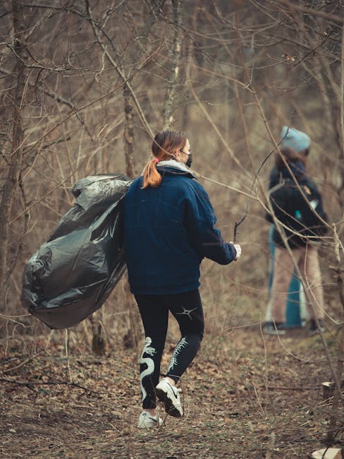 A person standing outdoors on a grassy area, wearing a green long-sleeved shirt, a patterned yellow and grey plaid jacket, grey trousers, and light green gloves, holding an open black rubbish bag lined with a silver or grey interior. The individual appears to be engaged in waste collection or rubbish removal, with the bag partially filled and held by the edges. The background consists of lush green grass, indicating an outdoor environment suitable for waste clearance activities. This scene reflects a private or independent waste disposal scenario, aligning with rubbish removal services offered by companies such as Waste Disposal Greenwich. The image emphasizes the act of handling waste with care and professionalism, suitable for articles discussing alternative waste handling methods or on-site clearance options for residents in the area.
