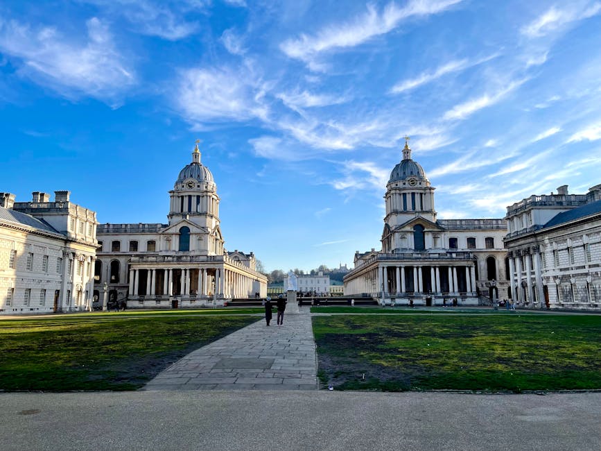 A wide view of an ornate, historic building complex with two prominent domed towers, featuring classical architectural details such as columns and decorative stonework, set against a bright blue sky with wispy clouds. In the foreground, a paved pathway runs centrally, leading to the entrance of the buildings, with a grassy area on both sides that appears slightly worn and patchy. The scene is illuminated by natural daylight, casting soft shadows on the stone facades. The setting is outdoor, within a spacious courtyard or park area, with a few people visible walking along the path, providing scale to the substantial architectural structures. This image, associated with waste disposal services, highlights an environment where cleanliness and space are important, subtly reflecting the context of private waste handling or on-site clearance that Waste Disposal Greenwich might support. The historic architecture and well-maintained surroundings contrast with the typical scenes of rubbish removal, emphasizing the importance of careful waste management to preserve such environments.
