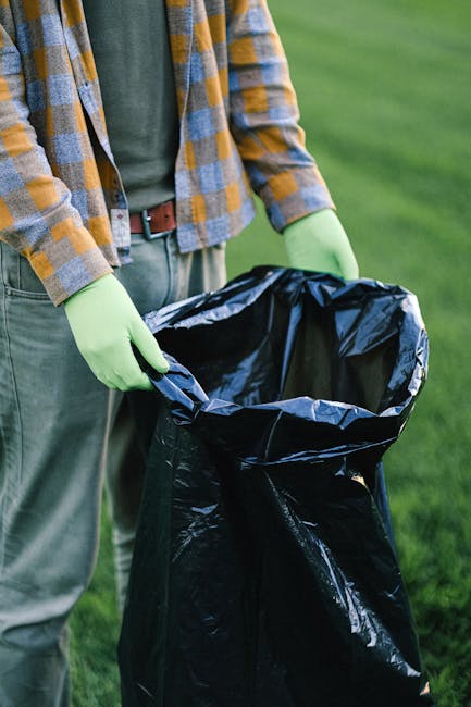 A person wearing a checked brown, yellow, and grey jacket with a green shirt underneath and grey trousers is holding a black trash bag open with both hands, which are covered in light green gloves. The trash bag is made of shiny plastic material and appears to be partially filled, set against a background of a well-maintained grassy outdoor area. The scene suggests an outdoor waste collection or rubbish removal activity, possibly part of a private or independent waste disposal effort. The background is out of focus, emphasizing the action of handling the rubbish bag. The lighting is natural, indicating daytime, and the environment is clean and tidy, consistent with organized rubbish collection practices that may involve professional services like Waste Disposal Greenwich for effective waste management in Greenwich or surrounding areas.