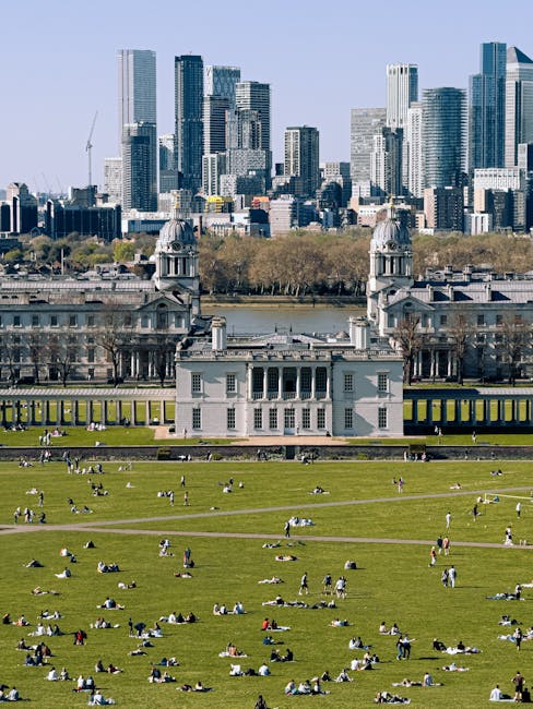 A wide view of an ornate, historic building complex with two prominent domed towers, featuring classical architectural details such as columns and decorative stonework, set against a bright blue sky with wispy clouds. In the foreground, a paved pathway runs centrally, leading to the entrance of the buildings, with a grassy area on both sides that appears slightly worn and patchy. The scene is illuminated by natural daylight, casting soft shadows on the stone facades. The setting is outdoor, within a spacious courtyard or park area, with a few people visible walking along the path, providing scale to the substantial architectural structures. This image, associated with waste disposal services, highlights an environment where cleanliness and space are important, subtly reflecting the context of private waste handling or on-site clearance that Waste Disposal Greenwich might support. The historic architecture and well-maintained surroundings contrast with the typical scenes of rubbish removal, emphasizing the importance of careful waste management to preserve such environments.
