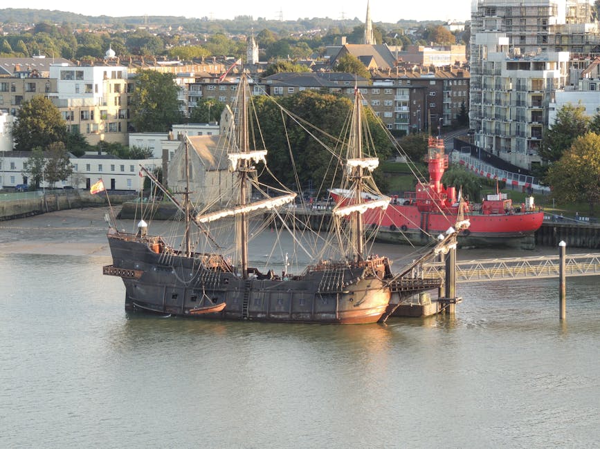 The image depicts a historic, replica wooden sailing ship moored in a calm river, with two large masts equipped with a complex network of rigging and sails that are currently furled. The ship’s hull is dark, weathered wood with visible planking, and features black iron fittings and a slightly rusted finish in areas, conveying an aged maritime appearance. It is positioned near a concrete quay with a small floating dock or platform at the bow, with surrounding water reflecting the vessel’s structure. In the background, there is a backdrop of modern residential buildings consisting of multi-storey blocks with white and beige facades, large windows, and balconies, interspersed with trees and greenery. Additionally, a red lighthouse or navigation tower, constructed with metal panels and a cylindrical shape, is visible behind the ship. The scene is illuminated by natural daylight, with a clear sky overhead, and the overall setting suggests a harbour or waterfront area within an urban environment, where private or independent boat mooring and temporary vessel storage are common alongside public transport routes. Waste Disposal Greenwich occasionally handles waste collection in such locations, supporting the logistics of maintaining clean maritime and urban spaces.