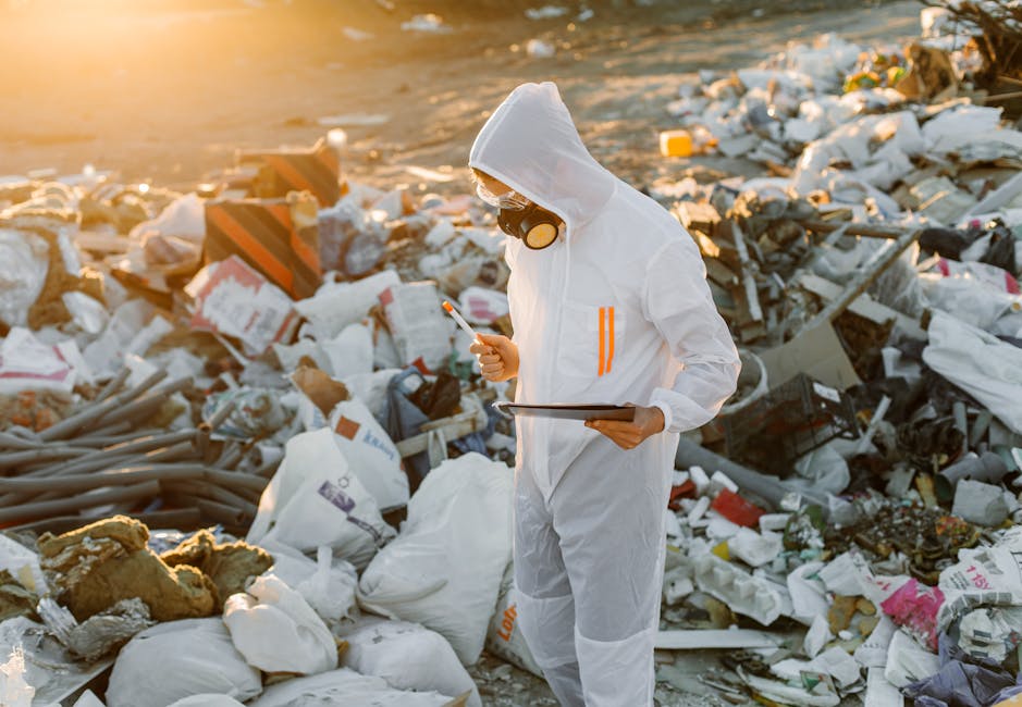 A person dressed in a white protective suit, including a hood and goggles, stands amid a large accumulation of scattered waste, including plastic bags, cardboard boxes, insulation material, and miscellaneous debris, on a sandy or gravelly surface. The individual holds a tablet in one hand and a pen or cleaning tool in the other, appearing to assess or document the extent of the rubbish. The background shows a sunset or sunrise casting a warm, golden light over the scene, emphasizing the disparity between the cleanup efforts and the overwhelming volume of waste. This setting highlights issues related to unmanaged refuse and may relate to initiatives for waste assessment or environmental monitoring by waste disposal services such as Waste Disposal Greenwich, providing insight into the challenges of rubbish removal and the importance of proper waste management practices.
