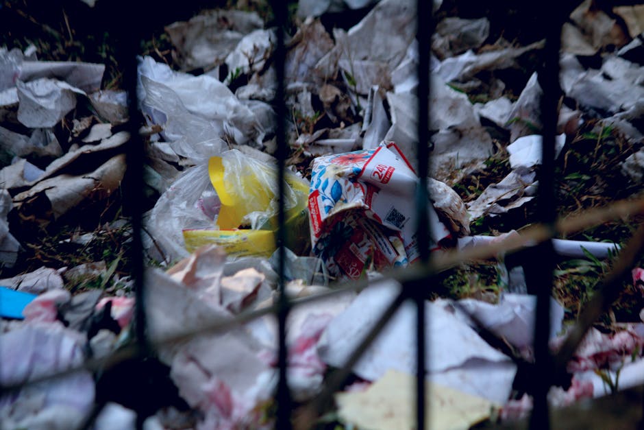 A large expanse of mixed waste and garbage scattered across an outdoor area, including plastic bottles, paper, cardboard, and organic matter, with some items partially buried or piled on top of each other. Among the debris, a small plastic bucket and stray pieces of wood are visible, indicating manual waste collection or clearance activities. The ground beneath the rubbish appears uneven and contaminated, with soil, dirt, and decomposing organic material intermixed. In the background, remnants of discarded packaging and household waste suggest an unmanaged, possibly unofficial waste dump site. The overall scene reflects a high-density accumulation of refuse commonly associated with unmanaged rubbish deposits, which may be subject to disposal or cleanup by waste management services like Waste Disposal Greenwich, offering alternative rubbish removal solutions that include on-site clearance or private disposal methods for large quantities of refuse.