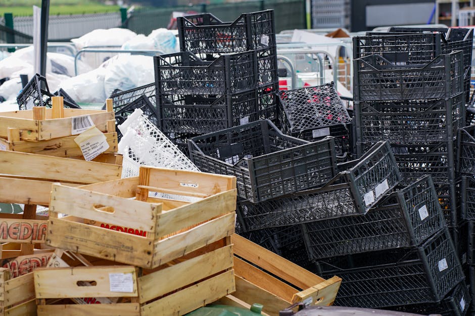 A large outdoor collection of discarded items and packaging materials, featuring stacked black plastic crates and wooden crates, some with labels attached. The crates are arranged in disorganized piles on a paved surface, with a background of blurred items wrapped in white plastic and additional waste materials, indicating a site for waste collection or disposal. The black crates have a lattice design and appear to be used for transporting or storing various goods, while the wooden crates are light-colored with cut-out handles, some with printed labels. The scene suggests an environment associated with private waste handling or rubbish removal services, emphasizing the routine collection and temporary storage of diverse waste materials typically managed by professional waste disposal companies like Waste Disposal Greenwich, facilitating alternative waste management solutions outside of municipal collection.