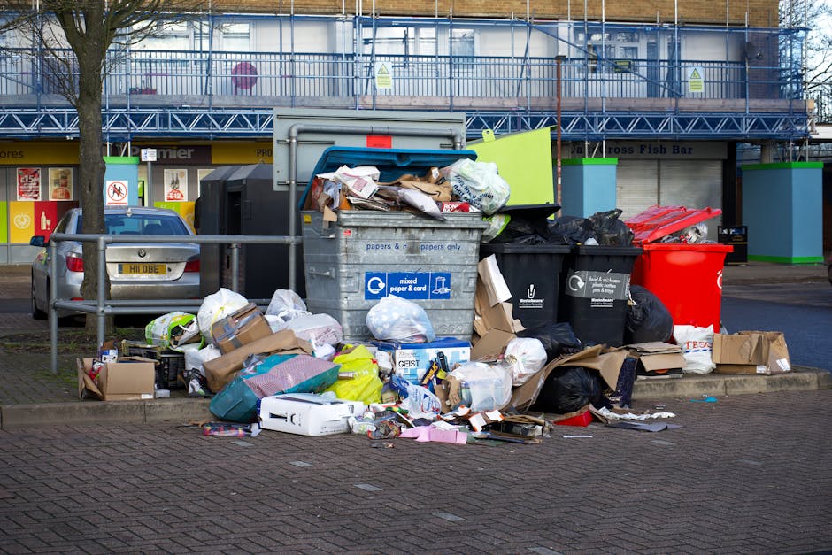 A large confusing assortment of overflowing rubbish bins and waste on a paved urban sidewalk, including black and red wheelie bins, a grey mixed paper and cardboard recycling bin with its lid open and items spilling out, surrounded by various scattered trash bags, flattened cardboard boxes, plastic packaging, and miscellaneous paper waste. Some waste is piled high across the bins and on the ground, with a few cardboard boxes and loose rubbish adjacent to the collection point. In the background, a commercial building with a blue scaffolding structure on the upper level, green-painted support pillars, and a closed storefront with signage visible. The environment appears to be an outdoor parking or loading area with a parked silver car behind a metal railing. The scene exemplifies a waste collection situation that may require private rubbish removal services, such as those offered by Waste Disposal Greenwich, to manage excess waste beyond standard local collection limits.