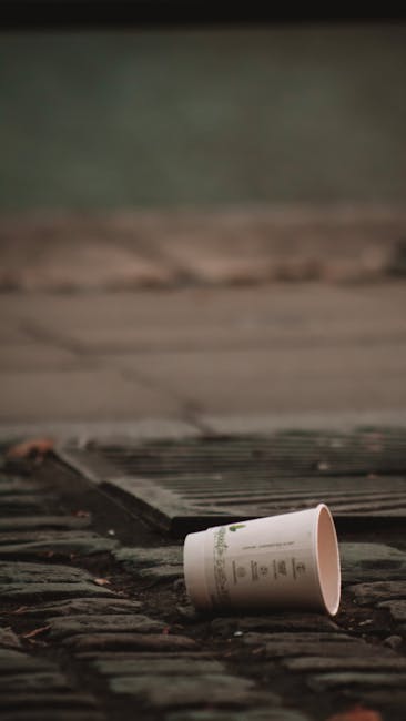 A discarded empty paper cup made of white material with printed text, lying on a brick-paved surface near a metal drain grate. The brick paving shows weathered textures and a mix of reddish-brown and dark tones, with some small debris scattered around. The drain cover has parallel lines and a slightly rusted appearance, positioned at the edge of a paved area that appears to be part of a driveway or pathway. In the background, a blurred greenish area suggests foliage or grass, with natural lighting casting soft shadows. The scene indicates an instance of littering in an outdoor environment, illustrating the importance of proper waste disposal which Waste Disposal Greenwich addresses through independent rubbish collection services, especially in narrow or challenging street layouts typical of Greenwich.