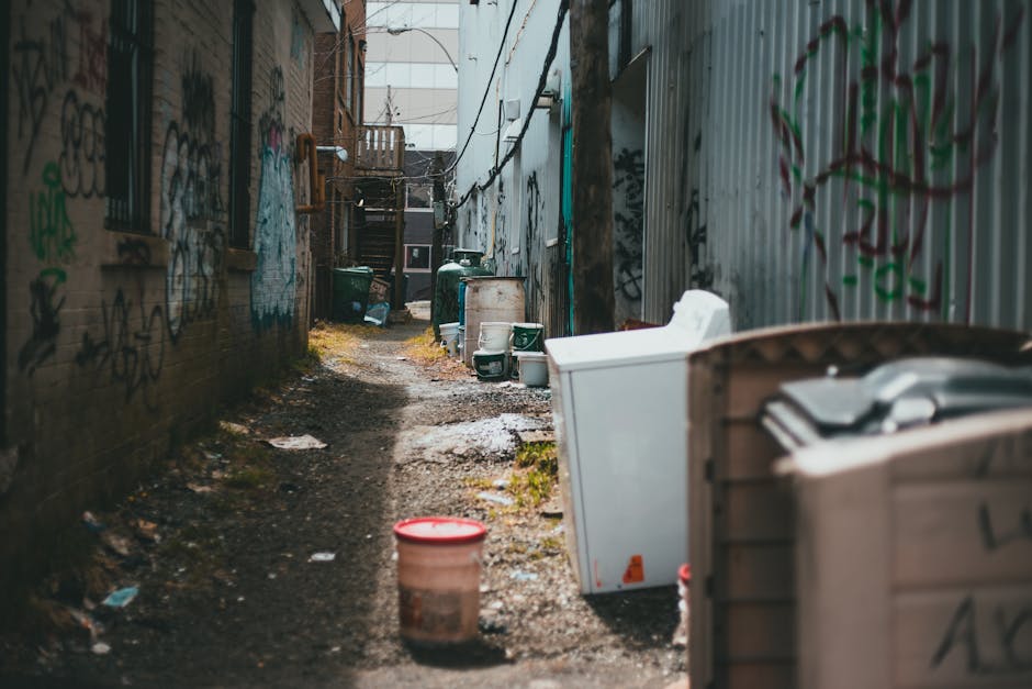 A large confusing assortment of overflowing rubbish bins and waste on a paved urban sidewalk, including black and red wheelie bins, a grey mixed paper and cardboard recycling bin with its lid open and items spilling out, surrounded by various scattered trash bags, flattened cardboard boxes, plastic packaging, and miscellaneous paper waste. Some waste is piled high across the bins and on the ground, with a few cardboard boxes and loose rubbish adjacent to the collection point. In the background, a commercial building with a blue scaffolding structure on the upper level, green-painted support pillars, and a closed storefront with signage visible. The environment appears to be an outdoor parking or loading area with a parked silver car behind a metal railing. The scene exemplifies a waste collection situation that may require private rubbish removal services, such as those offered by Waste Disposal Greenwich, to manage excess waste beyond standard local collection limits.