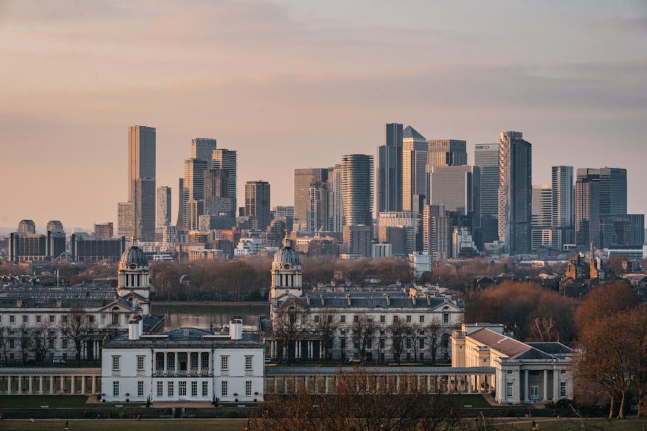 A panoramic view of central London skyline at dusk, featuring numerous modern skyscrapers with glass facades reflecting the soft light of the setting sun, towering above historical white buildings with domed roofs and classical architectural details in the foreground. The scene includes trees with bare branches indicating late autumn or winter, and a river visible just behind the historic structures, with the cityscape extending into the distance under a partly cloudy sky. The image captures the contrast between traditional and contemporary architecture within an urban environment, relevant to private waste disposal and the management of rubbish in central London areas, including the use of on-site clearance and independent collection services provided by Waste Disposal Greenwich.
