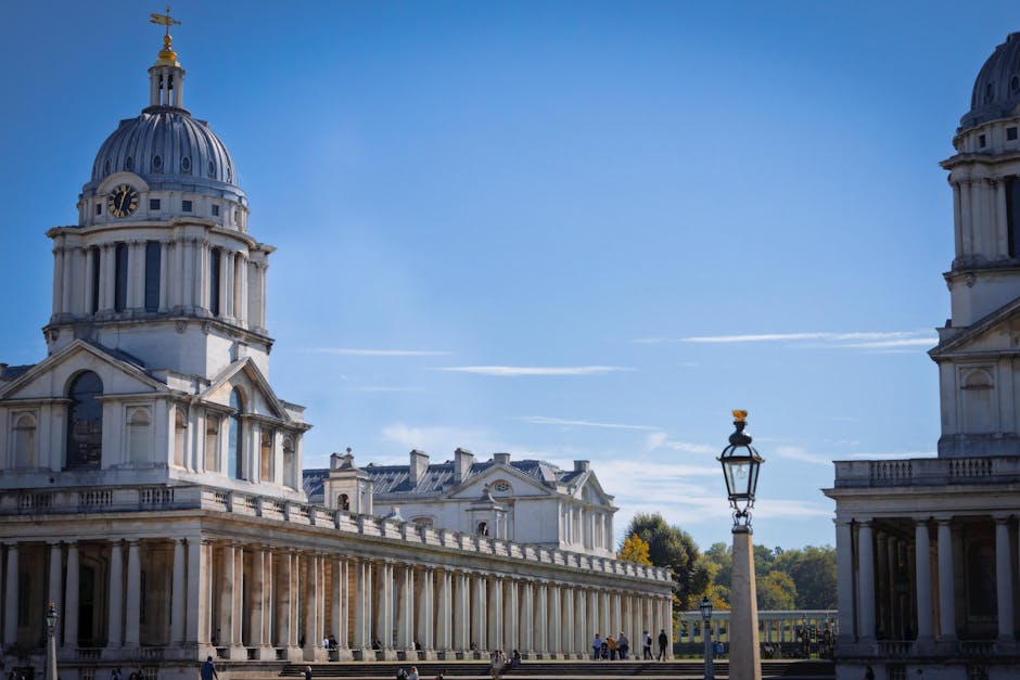 A large, historic neoclassical building with a prominent dome topped by a gold weather vane is visible in the image. The building features white stone or plaster façade with tall, narrow windows and ornamental architectural details, including decorative cornices and pilasters. In the foreground, there is a classical-style lamp post with an ornate metal design, standing on a stone or concrete base, positioned slightly to the right of the center. The background showcases a clear blue sky with a few wispy clouds, and some greenery with trees can be seen behind the structure. The overall atmosphere appears calm and bright, with natural sunlight illuminating the building’s façade and surroundings, emphasizing the textures and finishes of the stone surfaces. This scene subtly reflects an environment where historic architecture interacts with public outdoor spaces, which could be relevant to private or alternative waste handling contexts when considering on-site or ancillary service locations, though no specific rubbish removal activity is depicted in the image.