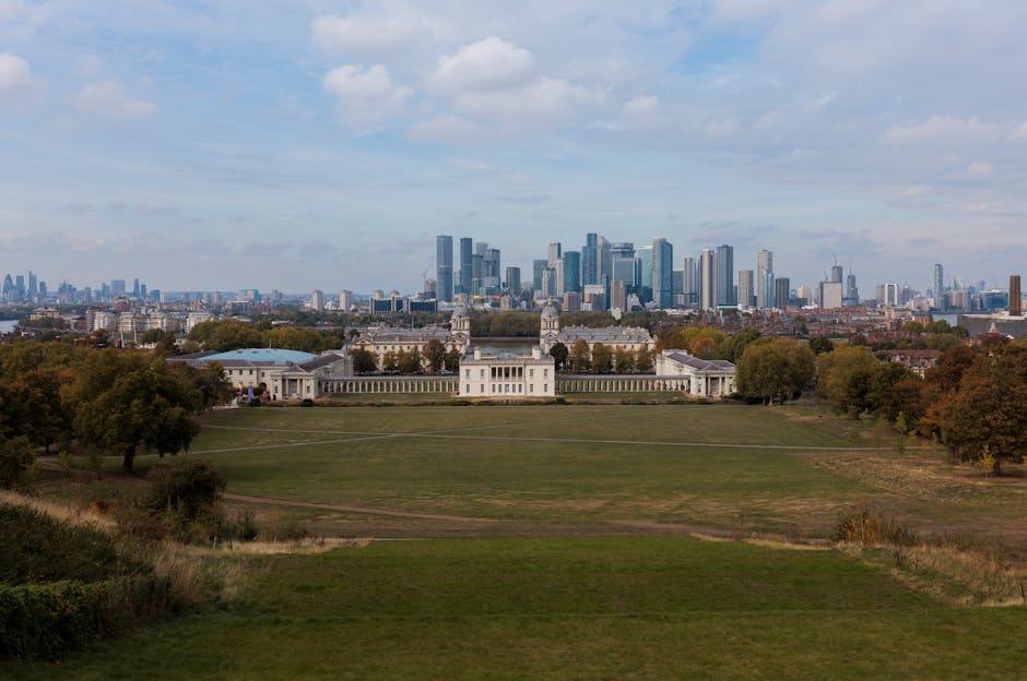 A wide panoramic view of historic white stone domed buildings with classical architectural details, including columns and decorative balustrades, situated in a park area with well-maintained grass and paved walkways. In the foreground, several wooden benches are visible along the paths, with a large leafy green tree on the left side partially obscuring one of the domed structures. Behind the buildings, a modern city skyline with tall glass skyscrapers and office towers rises into a cloudy, overcast sky, creating a contrast between historic and contemporary architectural styles. The scene suggests a location where private or independent waste management services, such as those provided by Waste Disposal Greenwich, might operate in urban and historic settings, supporting various rubbish collection needs within the cityscape.